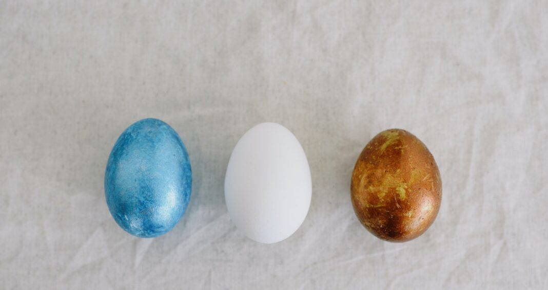 Three eggs (metallic blue, white and metallic gold) from left to right in a row on a white background.