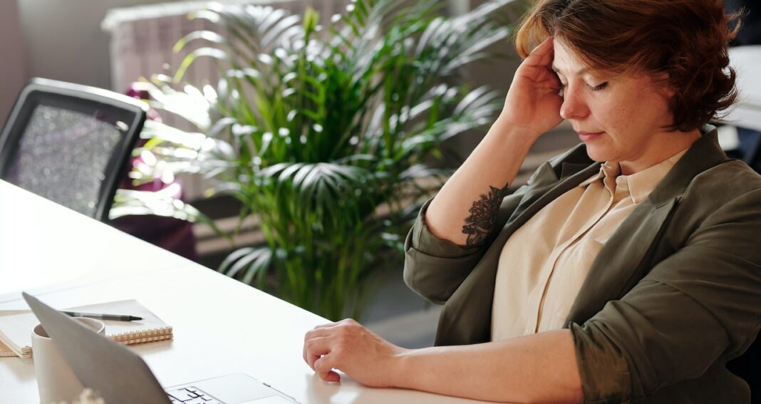 Woman sitting at desk in front of laptop holding her head with eyes closed.