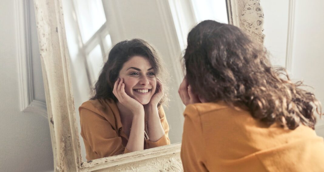 Woman in orange shirt looking at herself in a mirror.
