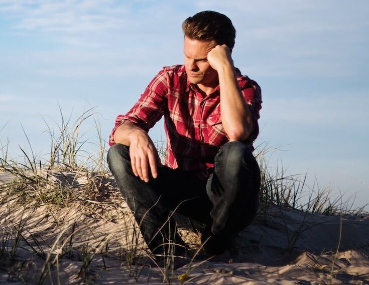 Man in red shirt sitting on a hill with fist on forehead thinking about something.