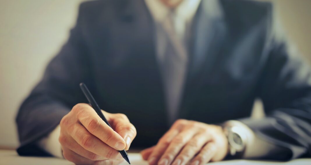 Hand with a pen writing something with a blurred in a suit behind from the neck down to waist.