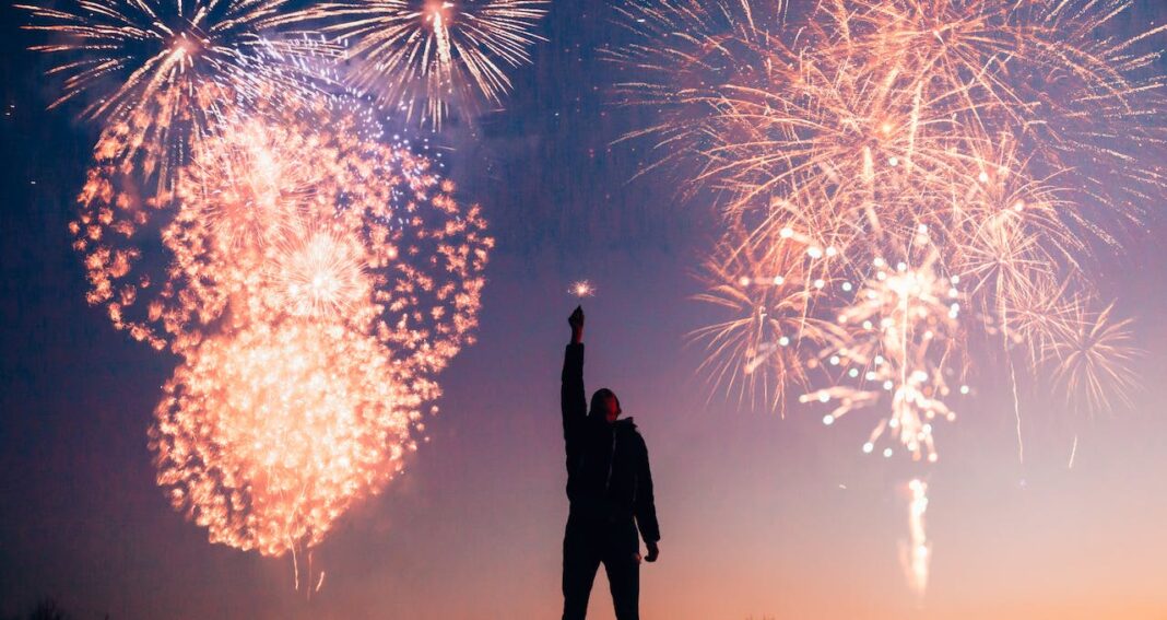 Man holding one arm high against a twilight sky with fireworks.